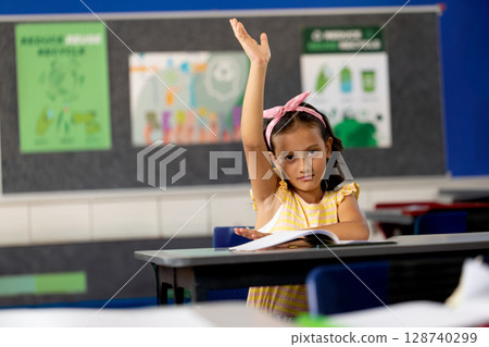 Female child raising hand at school desk in classroom with open notebook and colorful posters Female child raising hand at school desk in classroom with open notebook and colorful posters 128740299