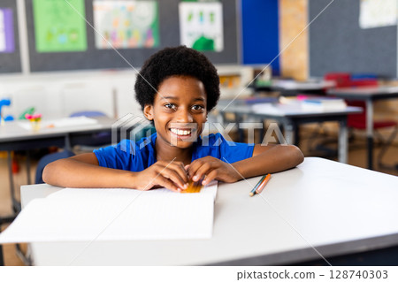 African American boy sitting at elementary classroom desk writing in open notebook with pencils African American boy sitting at elementary classroom desk writing in open notebook with pencils 128740303