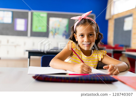 Child girl student writing in elementary classroom with blue wall, open notebook and red pencil 128740307