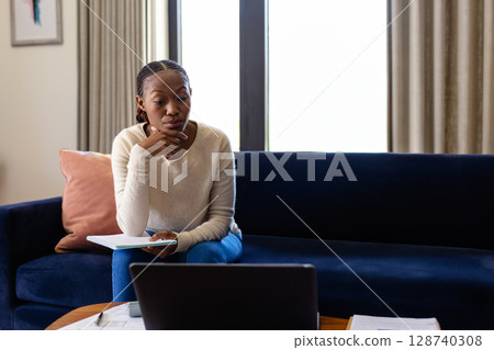 African American woman studying on sofa in living room, with laptop, notebook, pen, copy space African American woman studying on sofa in living room, with laptop, notebook, pen, copy space 128740308