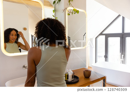 Young adult woman adjusting hair at bathroom vanity, with vine plant, lit mirror and soap bottles Young adult woman adjusting hair at bathroom vanity, with vine plant, lit mirror and soap bottles 128740309