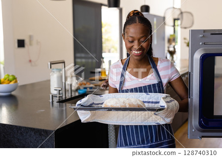 African American woman pulling tray from oven in modern kitchen, with loaf of bread, oven mitts African American woman pulling tray from oven in modern kitchen, with loaf of bread, oven mitts 128740310