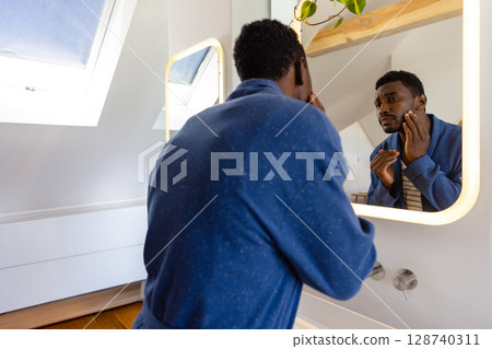 African American man examining face in attic bathroom with illuminated mirror, skylight and plant 128740311