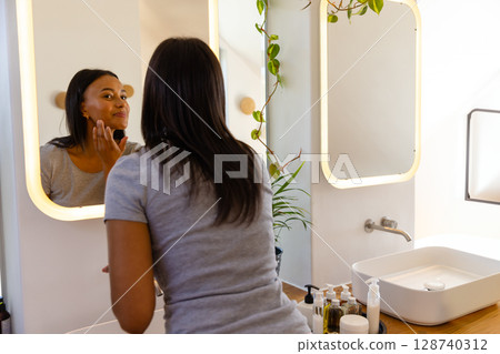 African American woman applying powder to cheek at bathroom vanity, holding black sponge 128740312
