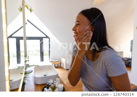 African American woman applying cream in attic bathroom by mirror, with skincare jars on counter 128740314