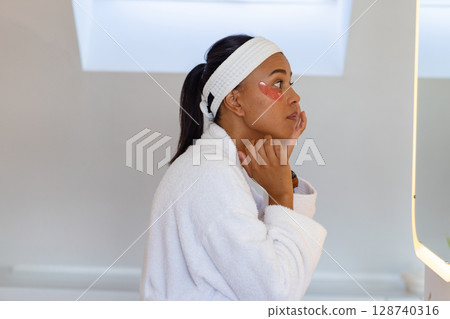 African American woman applying pink under-eye patch in bathroom, facing backlit mirror, copy space 128740316