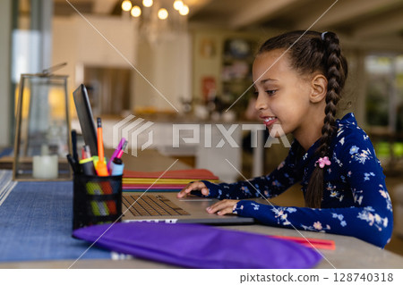 Child girl sitting at table using laptop in home kitchen, with pen holder, notebooks, copy space 128740318