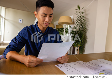 Asian man reading printed documents at wooden table in neutral home office, with potted plant Asian man reading printed documents at wooden table in neutral home office, with potted plant 128740320