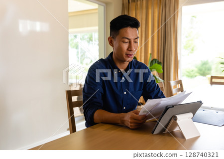 Young Asian man reviewing papers at wooden table in home office, with tablet and potted plant Young Asian man reviewing papers at wooden table in home office, with tablet and potted plant 128740321