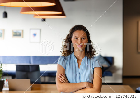 Woman standing confidently at table in office lounge, with pendant lamps, laptop and coffee cup 128740323
