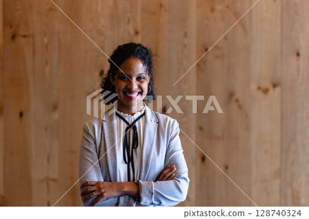 Young adult woman standing before wooden paneled wall in indoor studio, smiling with arms crossed 128740324