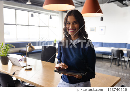 Woman standing at table in modern office, holding tablet beside laptop and brass lamp, copy space 128740326