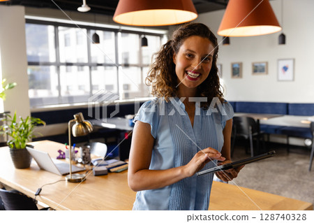 Female office professional using tablet in modern co-working space, with laptop desk lamp and plant 128740328