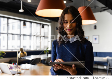 Woman tapping tablet in open plan office with laptop, pens in cup and plant, copy space 128740329