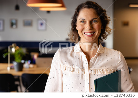 Woman standing and smiling while holding green folder in coworking office with wooden desk, laptop 128740330