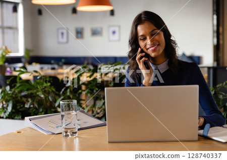 Woman talking on smartphone and working on laptop in office, with water and folder, copy space 128740337