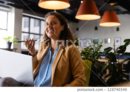 Young adult woman speaking on smartphone at desk in open-plan office lounge, with laptop and plants 128740340