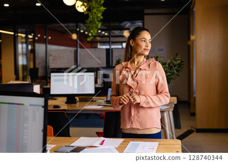 Businesswoman standing at wooden desk in office, using pink highlighter on papers, copy space 128740344
