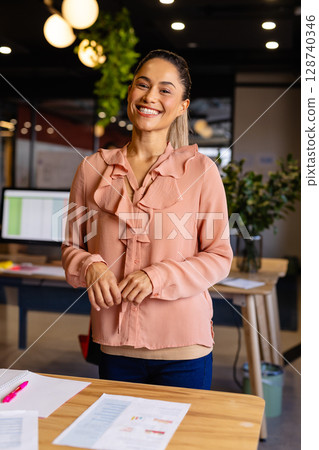 Woman in 30s standing behind wooden desk in modern office, smiling and reviewing charts 128740346