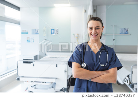 female nurse standing with arms crossed and smiling in hospital ward with IV stand, copy space 128740357