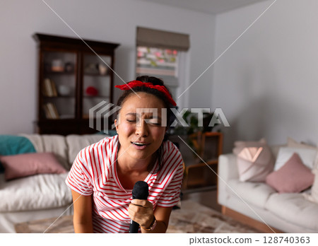 Chinese woman singing into handheld mic in cozy living room, showing wooden cabinet and pastel sofa Chinese woman singing into handheld mic in cozy living room, showing wooden cabinet and pastel sofa 128740363