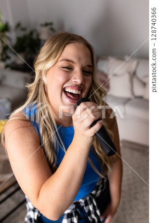 Young adult woman singing while standing in living room, holding handheld microphone and smiling 128740366