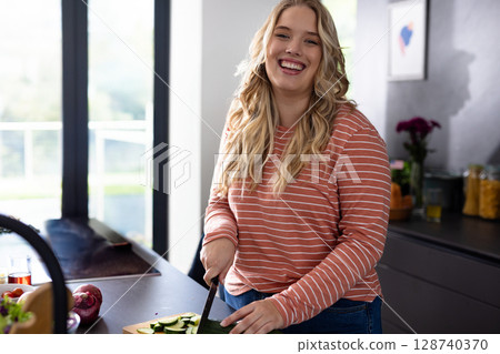 Young woman slicing cucumber in modern kitchen with kitchen knife on wooden cutting board 128740370