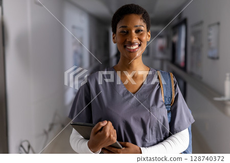African American woman standing in hospital corridor, holding tablet and wearing backpack, smiling 128740372