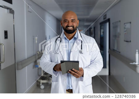 Male doctor holding tablet and smiling in hospital corridor, with stethoscope and IV stand 128740386