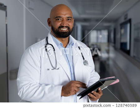 Male doctor using digital tablet in hospital hall wearing lab coat and stethoscope, copy space 128740389