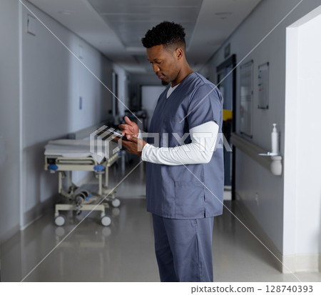 Young adult African American male healthcare worker tapping tablet in hospital corridor with gurney Young adult African American male healthcare worker tapping tablet in hospital corridor with gurney 128740393