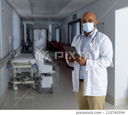 Male doctor reviewing tablet in hospital corridor, showing stethoscope and gurney, copy space 128740394