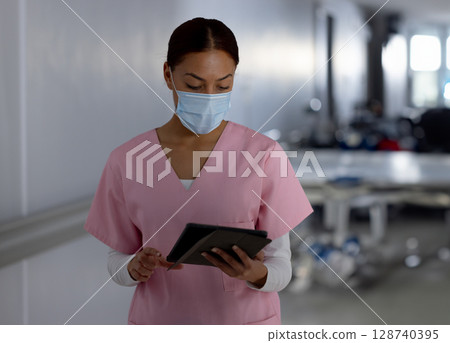 Female healthcare worker in pink scrubs studying tablet in hospital hallway wearing surgical mask 128740395