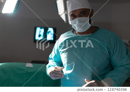 Male surgeon standing by draped table in dim operating room holding forceps with lights and monitor 128740396