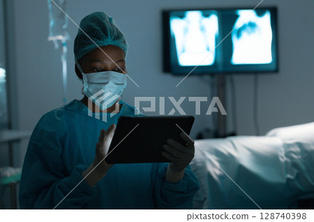 African American female surgeon reviewing tablet in dim hospital room with IV stand, copy space 128740398