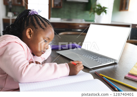 African American child writing at kitchen table with red pencil, open notebook, laptop, copy space African American child writing at kitchen table with red pencil, open notebook, laptop, copy space 128740401