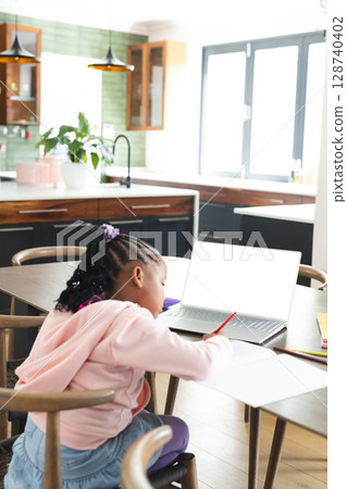 African American girl child leaning forward writing in notebook at kitchen table, with laptop African American girl child leaning forward writing in notebook at kitchen table, with laptop 128740402