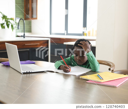African American boy writing in notebook at table in modern kitchen, with laptop and headphones African American boy writing in notebook at table in modern kitchen, with laptop and headphones 128740403