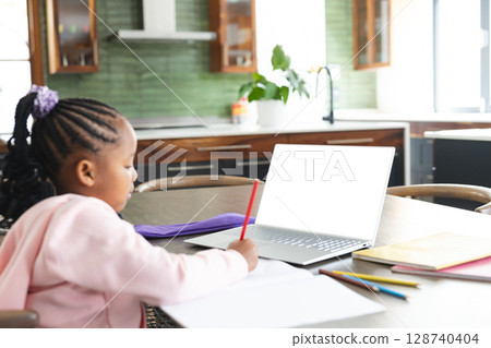 African American girl using silver laptop and writing with red pencil at kitchen table, copy space African American girl using silver laptop and writing with red pencil at kitchen table, copy space 128740404