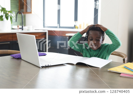African American boy sitting at kitchen table studying with headset, laptop, notebooks, copy space African American boy sitting at kitchen table studying with headset, laptop, notebooks, copy space 128740405