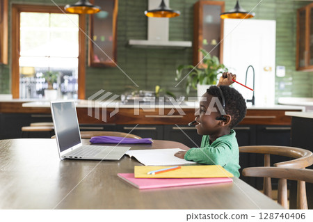 African American boy working on laptop in kitchen at table, with notebooks and headset, copy space 128740406