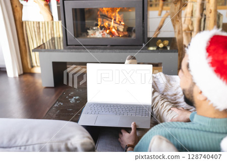 Man wearing Santa hat sitting on sofa at home holding laptop, with fireplace logs, copy space 128740407