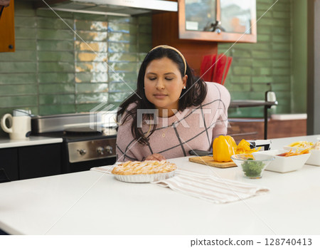 Woman leaning over kitchen island examining lattice pie in kitchen, with chopped peppers and cheese 128740413