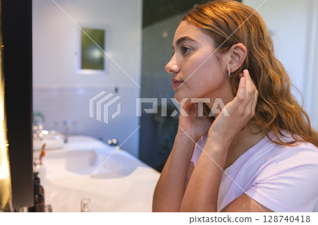 Woman adjusting earring and styling hair in home bathroom, with sink mirror and toiletry bottles Woman adjusting earring and styling hair in home bathroom, with sink mirror and toiletry bottles 128740418
