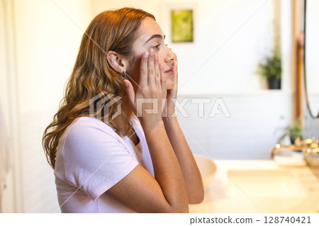 Woman applying skincare to cheeks at home bathroom vanity with modern faucet and potted plant Woman applying skincare to cheeks at home bathroom vanity with modern faucet and potted plant 128740421