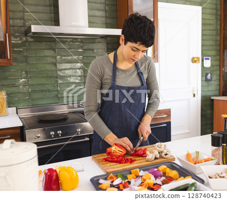 woman chopping red bell peppers on wooden cutting board in home kitchen, with spice jars 128740423