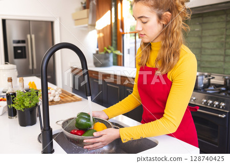 Young woman rinsing bell peppers in colander under gooseneck faucet in modern kitchen, copy space 128740425