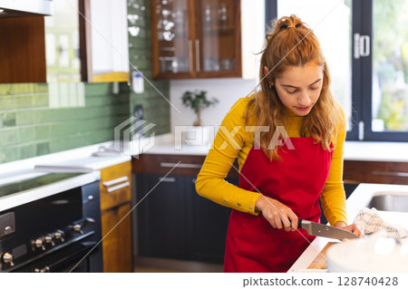 Young woman chopping vegetables on wooden cutting board in modern kitchen, with chef's knife Young woman chopping vegetables on wooden cutting board in modern kitchen, with chef's knife 128740428