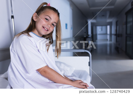 Female child sitting on hospital bed in hospital corridor, wearing gown with metal rails and pillow 128740429