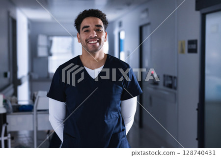 Young adult male healthcare worker standing in hospital corridor, smiling with medical cart behind 128740431